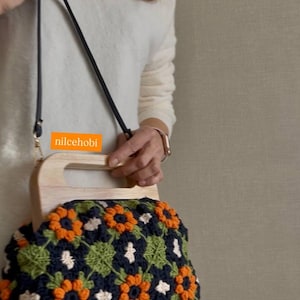 Puede incluir: Bolso de mano de ganchillo con asa de madera y correa negra. El bolso presenta un estampado floral en azul marino, naranja, verde y blanco. El texto "nilcehobi" es visible en el bolso.