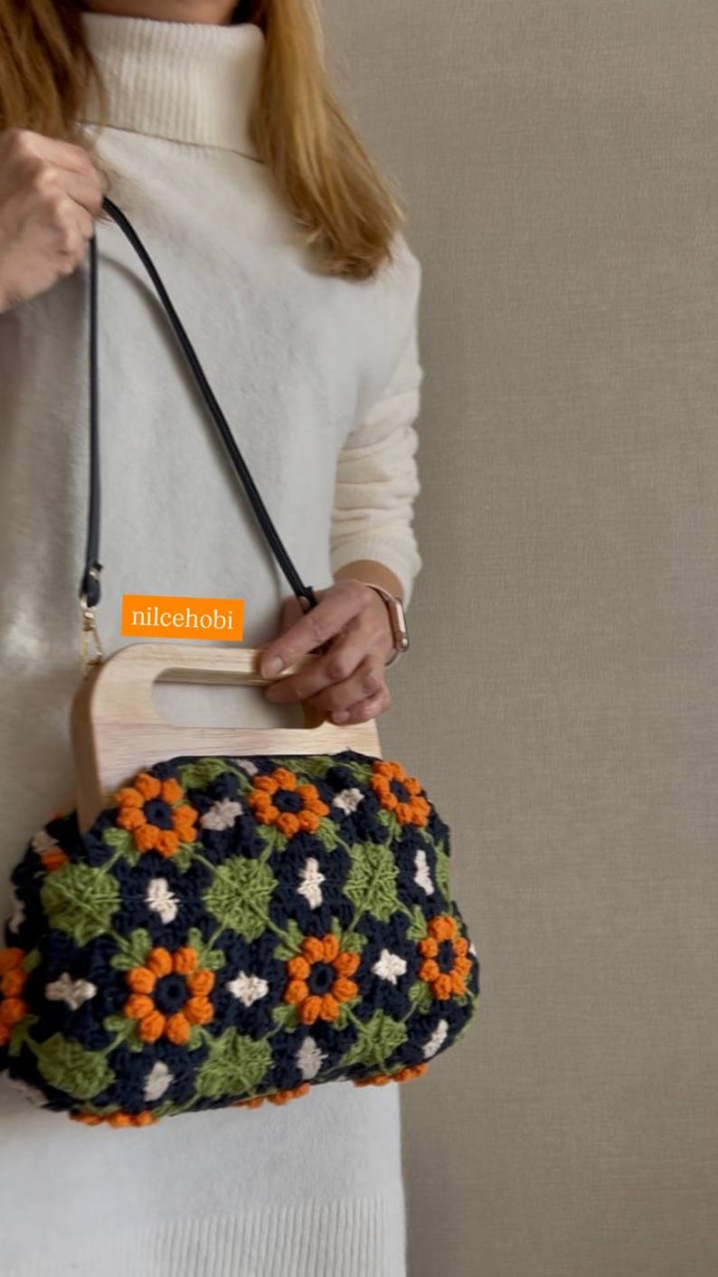 Puede incluir: Bolso de mano de ganchillo con asa de madera y correa negra. El bolso presenta un estampado floral en azul marino, naranja, verde y blanco. El texto "nilcehobi" es visible en el bolso.