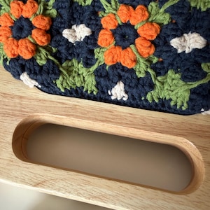Puede incluir: Primer plano de un bolso de ganchillo hecho a mano con un asa de madera. El bolso presenta un estampado floral en azul marino, naranja, verde y blanco. El asa de madera es de color marr&oacute;n claro y tiene un dise&ntilde;o curvo.