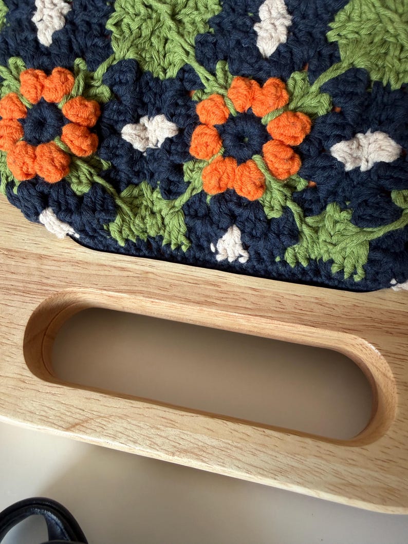Puede incluir: Primer plano de un bolso de ganchillo hecho a mano con un asa de madera. El bolso presenta un estampado floral en azul marino, naranja, verde y blanco. El asa de madera es de color marr&oacute;n claro y tiene un dise&ntilde;o curvo.