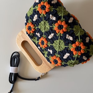 Puede incluir: Bolso de mano de ganchillo con asa de madera y correa negra desmontable. El bolso presenta un estampado floral con detalles en naranja, blanco y verde sobre un fondo azul marino. El asa de madera es rectangular con un recorte.