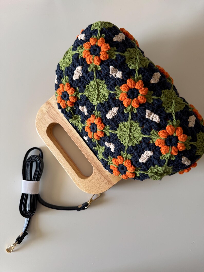 Puede incluir: Bolso de mano de ganchillo con asa de madera y correa negra desmontable. El bolso presenta un estampado floral con detalles en naranja, blanco y verde sobre un fondo azul marino. El asa de madera es rectangular con un recorte.