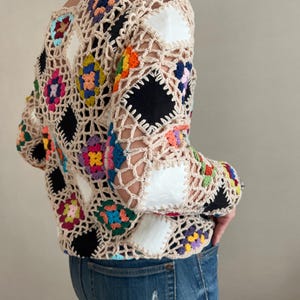 Peut inclure: Veste en crochet beige avec un motif patchwork. La veste présente des carrés colorés et des patchs noirs et blancs. Les manches sont longues et la veste est ouverte sur le devant. La veste est associée à un jean bleu.
