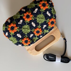 Puede incluir: Un bolso de mano de ganchillo con un asa de madera y una correa negra desmontable. El bolso presenta un estampado floral con detalles en naranja, verde y blanco sobre un fondo azul marino. El asa de madera tiene una abertura rectangular.