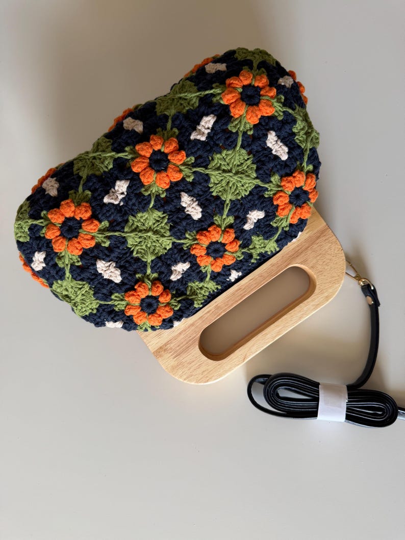 Puede incluir: Un bolso de mano de ganchillo con un asa de madera y una correa negra desmontable. El bolso presenta un estampado floral con detalles en naranja, verde y blanco sobre un fondo azul marino. El asa de madera tiene una abertura rectangular.