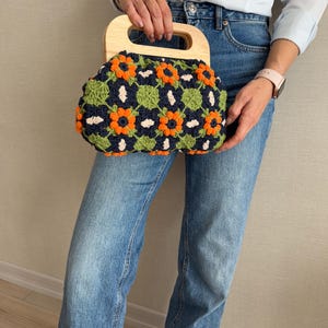 Puede incluir: Un bolso de mano de ganchillo con asa de madera. El bolso presenta un fondo azul marino con patrones florales naranjas y verdes. El asa es de madera clara.