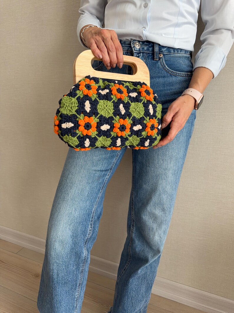 Puede incluir: Un bolso de mano de ganchillo con asa de madera. El bolso presenta un fondo azul marino con patrones florales naranjas y verdes. El asa es de madera clara.