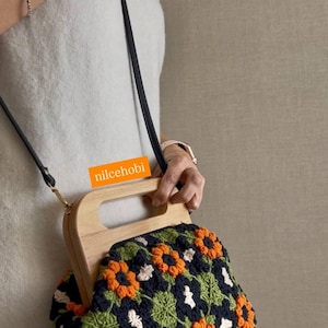 Puede incluir: Bolso de mano de ganchillo con asa de madera y correa para el hombro negra. El bolso presenta un estampado floral en naranja, verde, blanco y azul marino. El texto "nilcehobi" es visible en el asa.