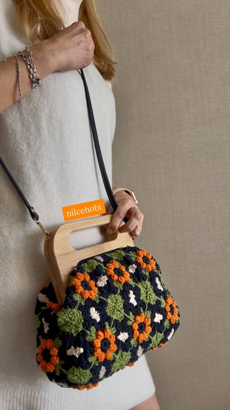 Puede incluir: Bolso de mano de ganchillo con asa de madera y correa para el hombro negra. El bolso presenta un estampado floral en naranja, verde, blanco y azul marino. El texto "nilcehobi" es visible en el asa.