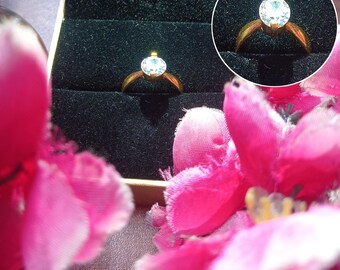 May include: A gold ring with a clear round gemstone in a black velvet ring box. The box is surrounded by pink fabric flowers.