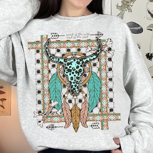 May include: A light gray sweatshirt featuring a turquoise and black cow skull design with feathers and arrows. The text "spirit of the wild soul of the west" is above the skull. The design is framed by a geometric pattern.
