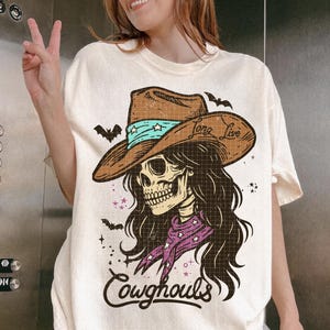 May include: Cream-colored t-shirt featuring a graphic of a skull wearing a cowboy hat with the text "Long Live" and "Cowghouls". The hat is brown with a turquoise band. Bats and stars are also included in the design.