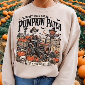 May include: A beige crewneck sweatshirt featuring a graphic print of a skeleton driving a tractor in a pumpkin patch. The text reads Support Your Local Pumpkin Patch and Best In Town Since 1922, accompanied by a scarecrow, bats, and pumpkins.