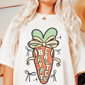 May include: A white t-shirt with a cartoon carrot design. The carrot is tied with a yellow ribbon and has the words "Happy Easter" written on it.