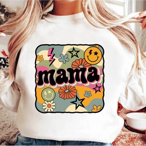 Op de afbeelding: Witte trui met een retro-grafisch ontwerp met het woord "mama" in een kleurrijk, golvend lettertype. Het ontwerp bevat bloemen, sterren en een smiley.