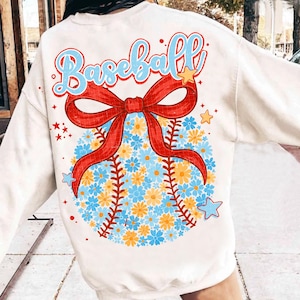 Puede incluir: Sudadera de color crema con un diseño de béisbol. La palabra "Baseball" está escrita en cursiva azul con detalles en rojo. Un lazo rojo está sobre una pelota de béisbol hecha de flores azules y amarillas. Estrellas dispersas alrededor del diseño.