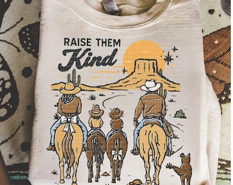 Może przedstawiać: Kremowa bluza z nadrukiem rodziny kowbojów na koniach w pustynnym krajobrazie. Tekst "RAISE THEM Kind" znajduje się nad ilustracją. Projekt zawiera słońce, kaktus i psa.