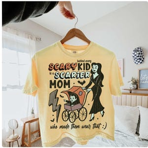 May include: Yellow t-shirt with the text "Behind every scary kid is a scarier mom who made them wear that :)". The graphic features a vampire mom pushing a stroller with a vampire child.