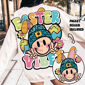 May include: Cream-colored sweatshirt with a colorful Easter-themed graphic. The design features a smiley face wearing a blue leopard-print beanie and bunny ears, with the words "EASTER VIBES" in bubble letters. Includes a pocket design.