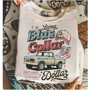 May include: Cream-colored sweatshirt with a graphic design featuring a blue and red vintage pickup truck, boots, and a baseball cap. Text includes "Living Blue Collar Dollar" and "Dirty Hands Clean Money."