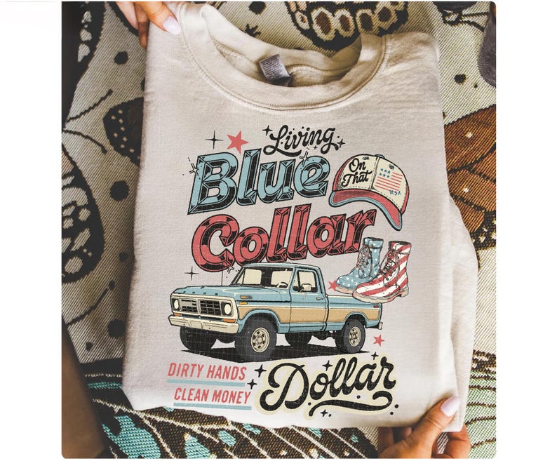 May include: Cream-colored sweatshirt with a graphic design featuring a blue and red vintage pickup truck, boots, and a baseball cap. Text includes "Living Blue Collar Dollar" and "Dirty Hands Clean Money."