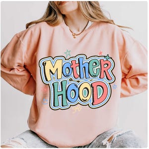 May include: A pale pink sweatshirt featuring the word "Motherhood" in a colourful, bubble-style font. The design includes stars, flowers, and a smiley face. The sweatshirt is long-sleeved with a crew neck.