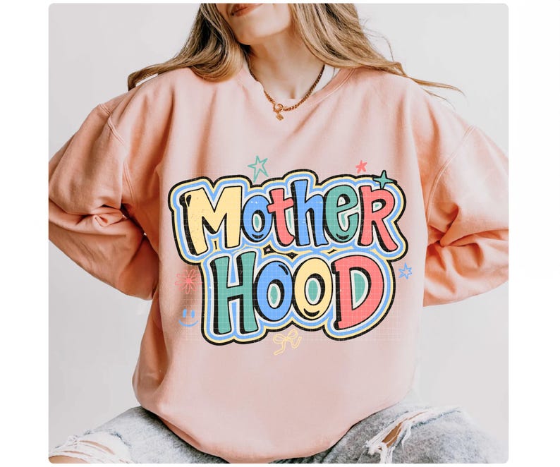 May include: A pale pink sweatshirt featuring the word "Motherhood" in a colourful, bubble-style font. The design includes stars, flowers, and a smiley face. The sweatshirt is long-sleeved with a crew neck.