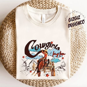 May include: Cream-colored t-shirt with a graphic design featuring a cowgirl riding a horse, a windmill, and the words "Cowgirls long love." The design includes red stars and blue clouds. The brand name "GIZGIZ DESIGNCO" is also visible.
