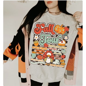 K&ouml;nnte beinhalten: Ein graues Sweatshirt mit einem bunten Grafikdruck, auf dem "Fall Tour" steht und Bilder von K&uuml;rbissen, Bl&auml;ttern und einem Truthahn zu sehen sind. Die Grafik enth&auml;lt auch Text wie "acorn findin", "apple pie bakin", "leaf rakin" und "scarf wearin".