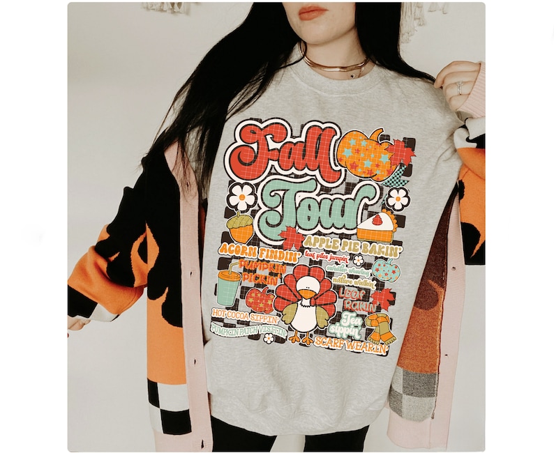 K&ouml;nnte beinhalten: Ein graues Sweatshirt mit einem bunten Grafikdruck, auf dem "Fall Tour" steht und Bilder von K&uuml;rbissen, Bl&auml;ttern und einem Truthahn zu sehen sind. Die Grafik enth&auml;lt auch Text wie "acorn findin", "apple pie bakin", "leaf rakin" und "scarf wearin".