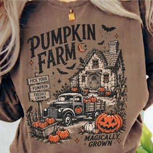 Peut inclure: Un sweat-shirt marron avec une impression graphique portant le texte PUMPKIN FARM et MAGICALLY GROWN. Le motif comprend une maison, un camion, des citrouilles, des chauves-souris et un panneau indiquant PICK YOUR PUMPKIN, PUMPKIN, FRESHLY GROWN.