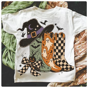 May include: White t-shirt featuring a Halloween-themed graphic. The design includes a witch's hat, cowboy boots with ghost and checkerboard patterns, bats, and a pumpkin. A checkered bow adds a decorative touch to the design.