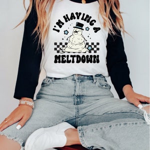 Pu&ograve; includere: Una maglietta da baseball bianca e nera con un grafico di pupazzo di neve che si scioglie e il testo "I'm having a meltdown".