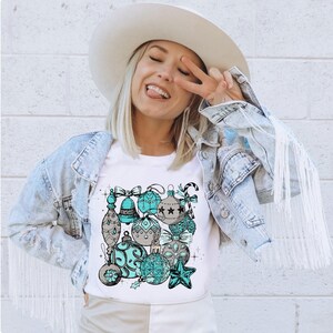 Peut inclure: Un t-shirt blanc avec un motif de boules de No&euml;l bleu sarcelle et grises. Les boules sont dispos&eacute;es en grappe et ont un style vintage.