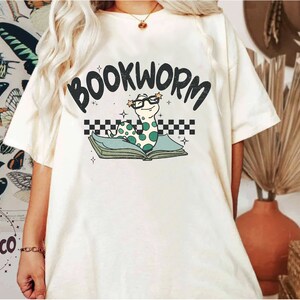 Pu&ograve; includere: Maglietta bianca con una grafica di un verme verde che indossa gli occhiali e legge un libro. Il testo "BOOKWORM" &egrave; sopra il verme.