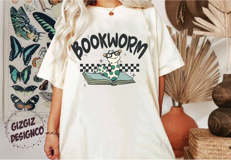 Pu&ograve; includere: Maglietta bianca con una grafica di un verme verde che indossa gli occhiali e legge un libro. Il testo "BOOKWORM" &egrave; sopra il verme.