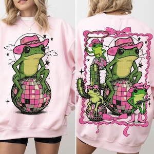 May include: Light pink sweatshirt featuring a green frog wearing a pink cowboy hat, sitting on a pink and silver disco ball. The back of the sweatshirt has a similar design with additional frogs, a cactus, and a pink frame.