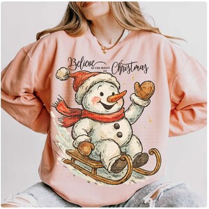 Op de afbeelding: Een lichtroze sweatshirt met een cartoon sneeuwpop op een slee. De sneeuwpop draagt een rode sjaal en hoed, en een wortelneus. De tekst luidt "Believe in the magic of Christmas".