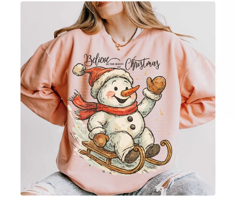 Op de afbeelding: Een lichtroze sweatshirt met een cartoon sneeuwpop op een slee. De sneeuwpop draagt een rode sjaal en hoed, en een wortelneus. De tekst luidt "Believe in the magic of Christmas".