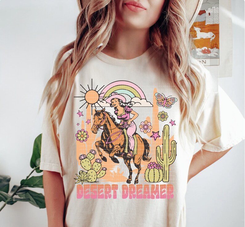 Pu&ograve; includere: Una maglietta bianca con un design grafico di una donna a cavallo in un paesaggio desertico. Il design include un arcobaleno, un cactus, stelle e il testo "Desert Dreamer".
