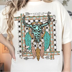 以下が含まれることがあります： クリーム色のTシャツ。 ターコイズと黒の雄牛の頭蓋骨のデザイン、羽、矢印が描かれています。 シャツには黒で「spirit of the wild soul of the west」と書かれています。 デザインには幾何学模様とアースカラーが使用されています。