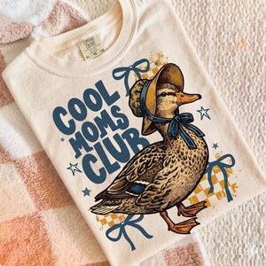 May include: Cream-colored t-shirt featuring a duck wearing a bonnet and a blue bow. The text "COOL MOMS CLUB" is printed in blue, with star accents. The shirt has a vintage, illustrated style.
