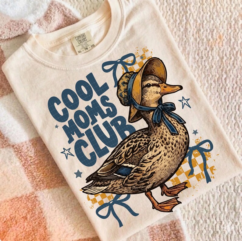 May include: Cream-colored t-shirt featuring a duck wearing a bonnet and a blue bow. The text "COOL MOMS CLUB" is printed in blue, with star accents. The shirt has a vintage, illustrated style.