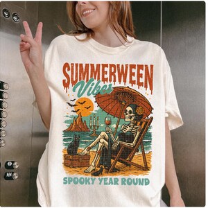 May include: Cream-colored t-shirt with a graphic featuring a skeleton relaxing on a beach chair under an umbrella, holding a glass of wine. The text reads "SUMMERWEEN Vibes" and "SPOOKY YEAR ROUND". A black cat sits nearby.