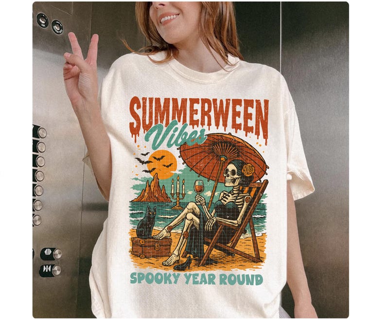 May include: Cream-colored t-shirt with a graphic featuring a skeleton relaxing on a beach chair under an umbrella, holding a glass of wine. The text reads "SUMMERWEEN Vibes" and "SPOOKY YEAR ROUND". A black cat sits nearby.