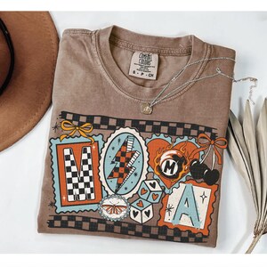 Op de afbeelding: Een bruin Comfort Colors T-shirt met een grafisch ontwerp met de letters "MAMA" in decoratieve kaders. Het ontwerp bevat een bliksemschicht, dobbelstenen, kersen en een vlinder. Een zilveren ketting met een hanger ligt op het shirt.