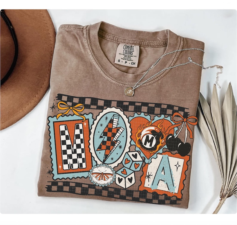 Op de afbeelding: Een bruin Comfort Colors T-shirt met een grafisch ontwerp met de letters "MAMA" in decoratieve kaders. Het ontwerp bevat een bliksemschicht, dobbelstenen, kersen en een vlinder. Een zilveren ketting met een hanger ligt op het shirt.