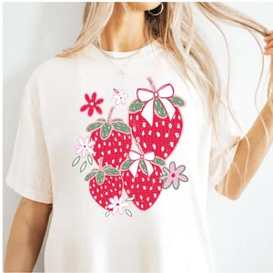 May include: White t-shirt with a graphic of red strawberries with green leaves and pink flowers. The strawberries have white polka dots and white bows.