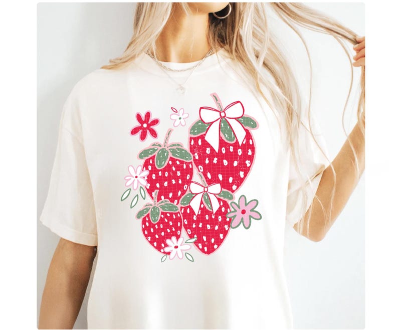 May include: White t-shirt with a graphic of red strawberries with green leaves and pink flowers. The strawberries have white polka dots and white bows.