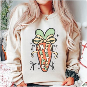 May include: A cream-colored sweatshirt with a cartoon carrot design. The carrot is decorated with a green bow and the words "Happy Easter" are written on the carrot.